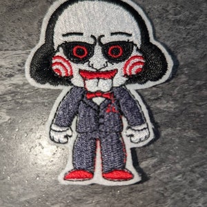 May include: An iron-on patch embroidered with a cartoon character. The character has a white face, red lips, and black hair. They are wearing a black suit, red bow tie, and red and white gloves. The character's eyes are red and have a spiral pattern.