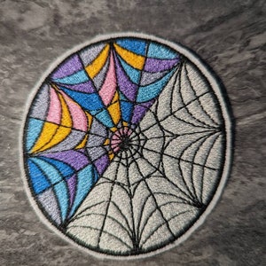May include: A circular embroidered patch featuring a colourful stained glass design on one half and a black and white spiderweb design on the other half.