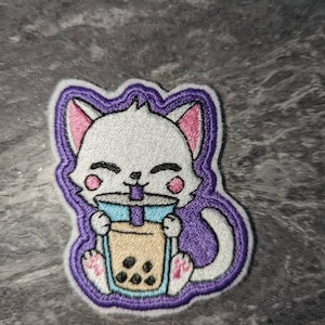 May include: White embroidered patch of a cat holding a cup of bubble tea with a purple outline.