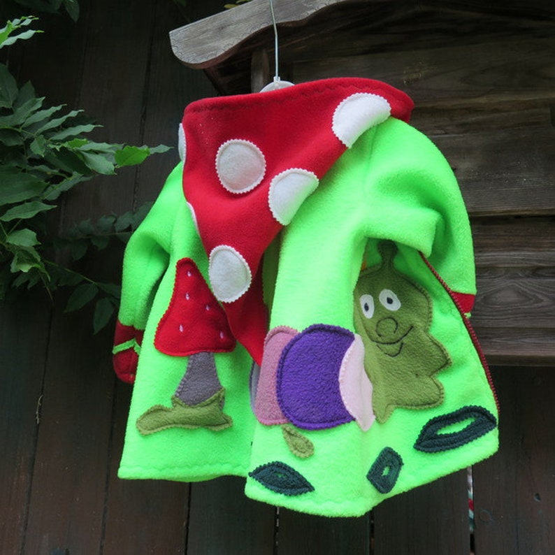 Puede incluir: Una chaqueta de forro polar verde con una capucha de hongo rojo y una aplicaci&oacute;n de caracol. La chaqueta tiene lunares blancos en la capucha y hojas verdes alrededor del caracol.