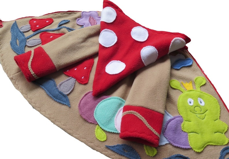 Puede incluir: Una chaqueta de forro polar de color beige con ribetes de forro polar rojo y aplicaciones de un hongo, fresas, hojas y una oruga verde con una corona.