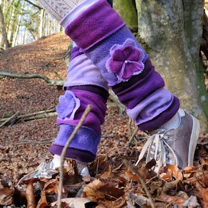 Könnte beinhalten: Lila Fleece-Beinwärmer mit Blumenverzierungen. Die Beinwärmer sind in verschiedenen Lilatönen gehalten und haben eine Blumenapplikation an jedem Bein. Die Person trägt braune Turnschuhe und steht auf Herbstlaub in einem Wald.