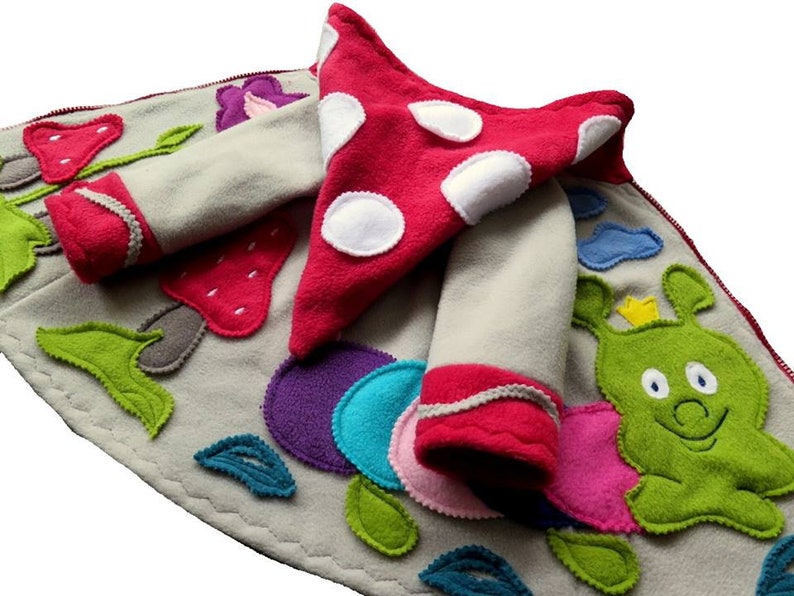 Puede incluir: Una chaqueta de forro polar gris con una capucha de hongo rojo y aplicaciones coloridas de flores, hojas y una oruga. La chaqueta tiene una cremallera y pu&ntilde;os rojos.