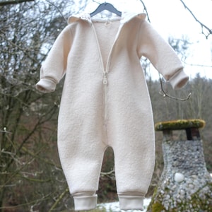 May include: Cream-colored baby jumpsuit with a hood and a full-length zipper. The one-piece garment has long sleeves and ribbed cuffs at the wrists and ankles. The soft, textured fabric is hanging on a black hanger against a blurred outdoor background.