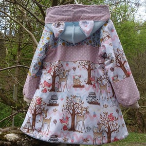 Könnte beinhalten: Eine rosa und blaue Kapuzenjacke mit einem weißen, rosa und braunen Waldtier-Druck. Die Jacke hat eine Kapuze mit zwei Ohren und einem weißen Futter. Die Jacke ist aus einem weichen, wasserabweisenden Stoff.