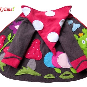 Puede incluir: Una chaqueta de forro polar marr&oacute;n con una capucha de hongo rojo, una oruga verde y aplicaciones de flores de colores. La chaqueta tiene una cremallera y un ribete rojo alrededor de las mangas y la capucha.