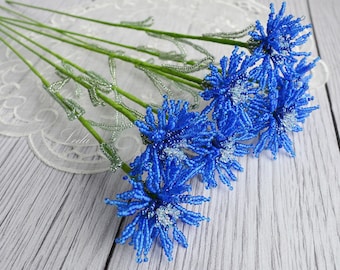 May include: A bouquet of blue beaded flowers with green stems. The flowers are arranged on a white wooden surface.