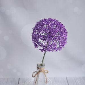 Peut inclure: Une sculpture florale sphérique composée de perles violettes avec une fine tige verte, placée dans une bouteille en verre transparent. La bouteille est ornée d'un nœud en ficelle brune noué autour du goulot, formant un ensemble décoratif.