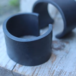 May include: Two black metal bracelets, one stacked on top of the other, with a smooth, matte finish.