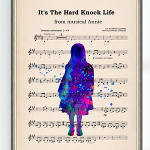 Könnte beinhalten: Gerahmter Druck mit Notenblättern und einer Aquarell-Silhouette einer Person. Die Silhouette ist mit leuchtenden Blau-, Lila- und Rosatönen gefüllt. Der Text oben lautet "It's The Hard Knock Life" aus dem Musical Annie.