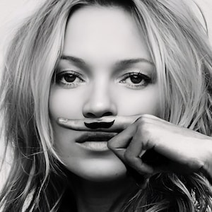 May include: Black and white portrait of a person with long, wavy hair. A finger is held up to the lips, with a drawn-on mustache. The image is a close-up, focusing on the face and the playful mustache.