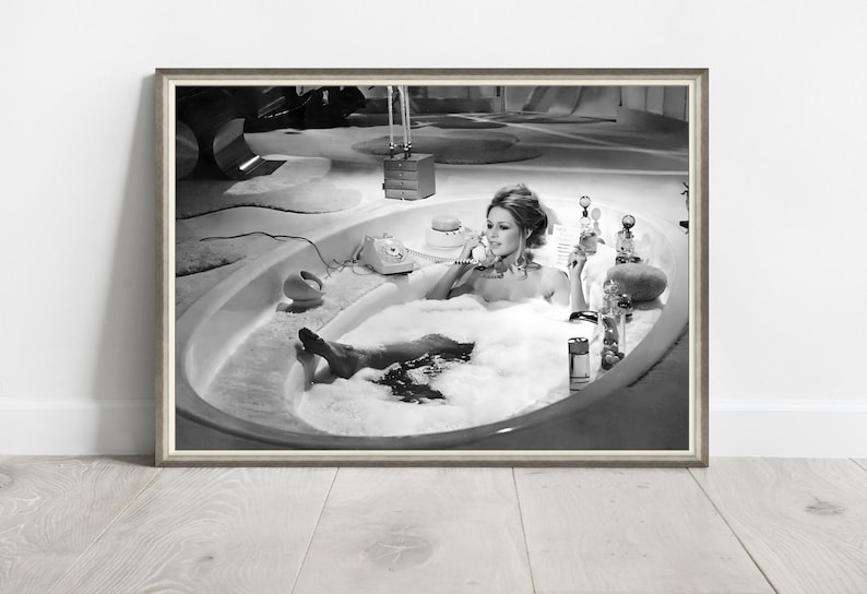 May include: A black and white photograph in a thin frame depicting a person with hair styled in an updo, reclining in an oval-shaped bathtub filled with white foam. The person is holding a telephone receiver to their ear, surrounded by various bottles.