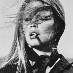 May include: Black and white portrait of a person with long, flowing hair and a cigarette in their mouth. The person is wearing a dark top and scarf. The image is a classic, vintage style.