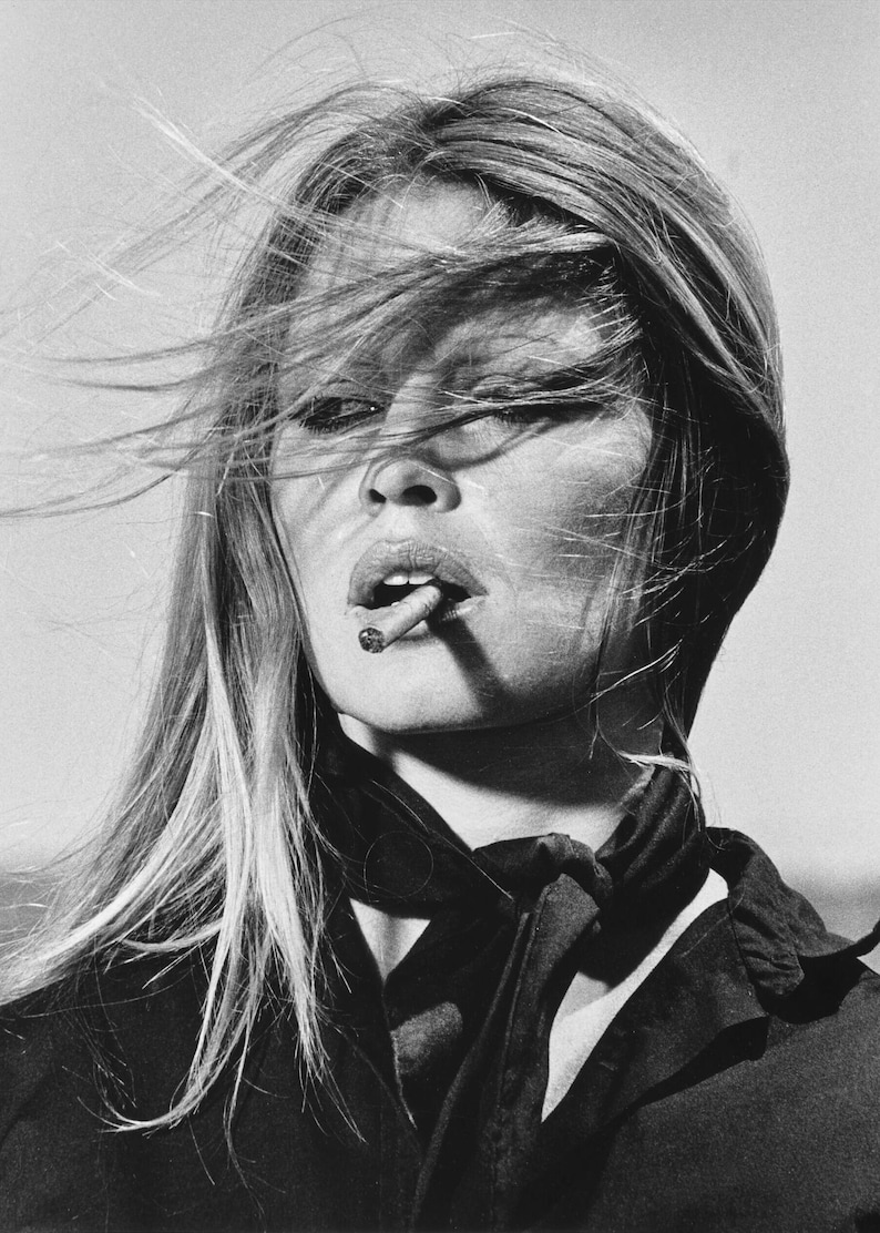May include: Black and white portrait of a person with long, flowing hair and a cigarette in their mouth. The person is wearing a dark top and scarf. The image is a classic, vintage style.