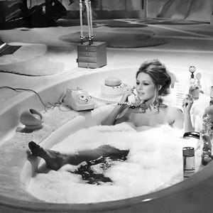 May include: A black and white photograph of a person in a large oval bathtub filled with white foam. The person is holding a vintage rotary telephone handset to their ear. Various glass perfume bottles and a small round container sit on the tub edge.