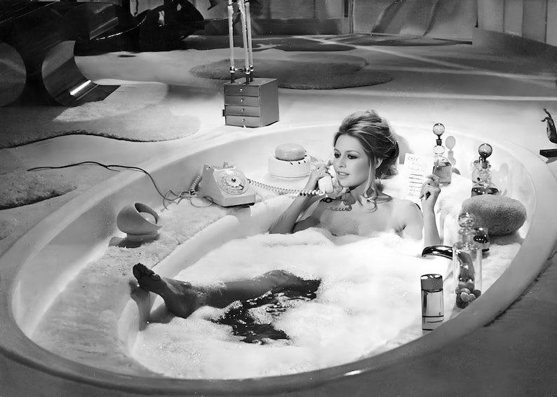 May include: A black and white photograph of a person in a large oval bathtub filled with white foam. The person is holding a vintage rotary telephone handset to their ear. Various glass perfume bottles and a small round container sit on the tub edge.