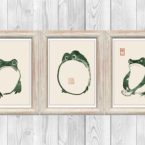 Peut inclure: Trois impressions encadrées de grenouilles vertes dans différentes poses. La première grenouille est assise la bouche ouverte. La deuxième grenouille est enroulée en cercle. La troisième grenouille est assise avec un bol devant elle.