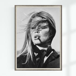 May include: Black and white framed print of a woman with long, wind-blown hair, smoking a cigarette. She wears a dark top and scarf. The portrait is a classic, vintage style.