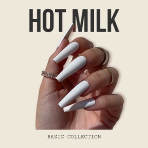 Puede incluir: Uñas postizas blancas, largas, en forma de ataúd. La imagen incluye el texto "HOT MILK" en la parte superior y "BASIC COLLECTION" en la parte inferior. La mano está adornada con anillos dorados y un anillo con forma de diamante.