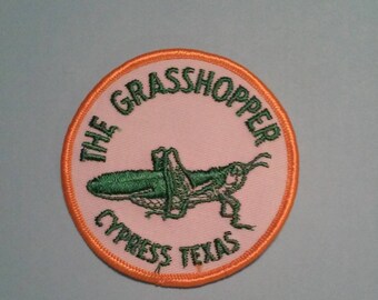May include: A round, orange-bordered patch with a pink background. The patch features a green embroidered grasshopper and the text "THE GRASSHOPPER CYPRESS TEXAS".