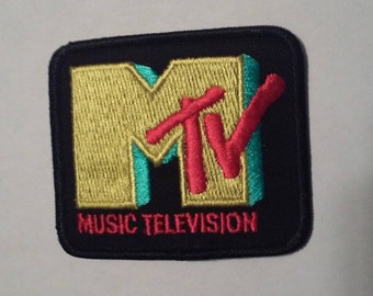 May include: A black embroidered patch with the MTV logo in yellow, red, and teal. The text "MUSIC TELEVISION" is below the logo.