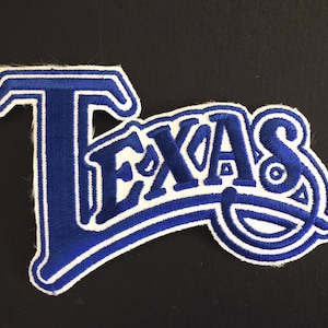 May include: Blue and white embroidered patch with the word "TEXAS" in a white outline. The patch is shaped like a curved letter "T" with the word "TEXAS" inside the curve.