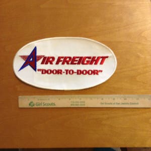 May include: White oval patch with red embroidered text "AIR FREIGHT" and "DOOR-TO-DOOR" with a blue and red star in the upper left corner.