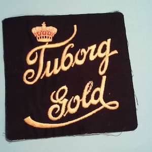 May include: A black fabric patch with gold embroidery. The patch features a crown and the words "Tuborg Gold".