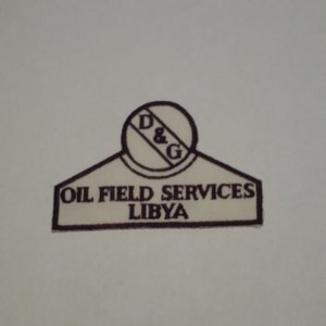 May include: A white patch with brown embroidered text and a circle. The text reads "OIL FIELD SERVICES LIBYA" and the circle has the letters "D & G" inside.