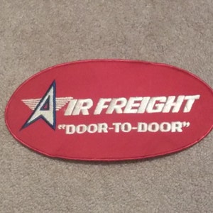 May include: Red oval patch with a white embroidered airplane logo and the text "Air Freight" in white with the tagline "Door-to-Door" in white.