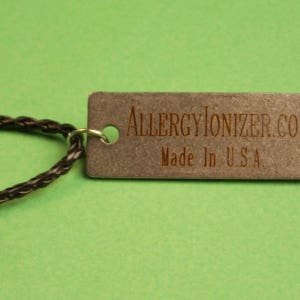 May include: A close-up of a silver tag with the text "AllergyIonizer.com" and "Made in U.S.A." The tag is attached to a black braided cord.