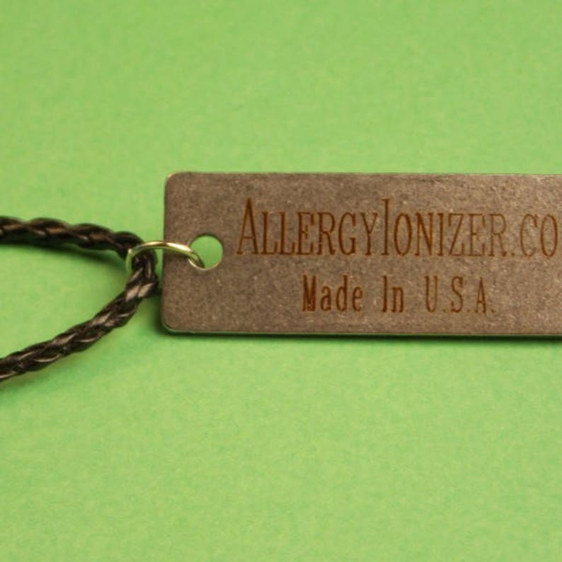 May include: A close-up of a silver tag with the text "AllergyIonizer.com" and "Made in U.S.A." The tag is attached to a black braided cord.