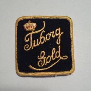 May include: A black fabric patch with gold embroidered lettering that reads "Tuborg Gold". A gold crown is above the text.