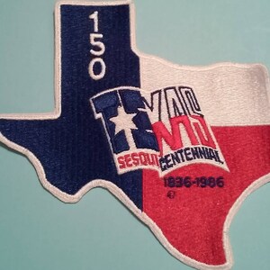 May include: A blue and red embroidered patch shaped like the state of Texas. The patch features the text "150" in the top left corner, "Texas" in the center, "Sesquicentennial" and "1836-1986" in the bottom center.