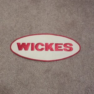 Puede incluir: Parche ovalado blanco con un borde rojo bordado y la palabra "WICKES" en rojo.