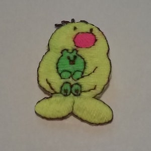 May include: A yellow felt applique of a bird holding a green felt applique of a frog in its arms. The bird has a pink felt nose and black felt eyes.