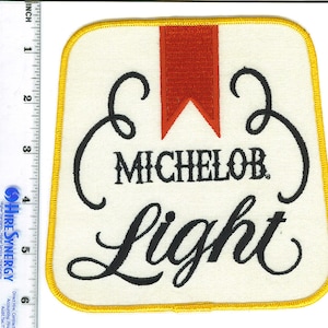 May include: A white embroidered patch with a yellow border. The patch features a red ribbon and the text "MICHELOB Light" in a black cursive font.