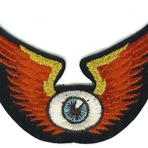 May include: Embroidered patch featuring a stylized eye with orange and yellow wings. The eye has a blue iris and white sclera, set against a black background. The wings are detailed with orange and yellow thread.
