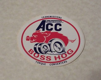 May include: Oval sticker featuring a red cartoon pig driving on wheels, with the text "ACC Performance" and "Boss Hog Torque Converters" in red and blue. The sticker has a white background and a red and blue border.