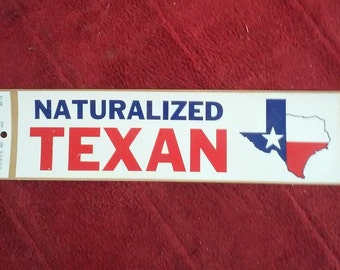 May include: A white sticker with the text "NATURALIZED TEXAN" in blue and red letters. A blue and red outline of the state of Texas with a white star is on the right side of the sticker.