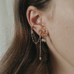May include: Close-up of an ear featuring multiple gold earrings. The earrings include a chain, a star-shaped stud, and a hoop with dangling crystals. The jewellery is set against a neutral backdrop, highlighting the intricate details of the pieces.