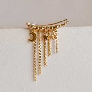 Może przedstawiać: Złota nausznica z księżycem w kształcie półksiężyca, gwiazdami i wiszącymi łańcuszkami. Nausznica jest ozdobiona małymi, okrągłymi koralikami i dopasowuje się do kształtu ucha. Biżuteria o tematyce niebiańskiej jest prezentowana na białym tle. Około 3 cm długości.