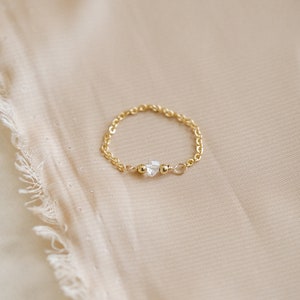 May include: A delicate gold chain ring with a clear crystal stone accent. The ring is on a light beige fabric background.
