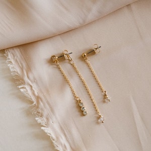 Op de afbeelding: Goudkleurige oorhangers met heldere kristallen accenten. Eén oorbel heeft drie kristallen, de andere één kristal. De oorbellen worden getoond tegen een beige stoffen achtergrond.