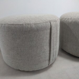 K&ouml;nnte beinhalten: Zwei runde graue Stoffhocker mit einer dezenten Textur. Die Hocker haben eine Naht an der Seite.