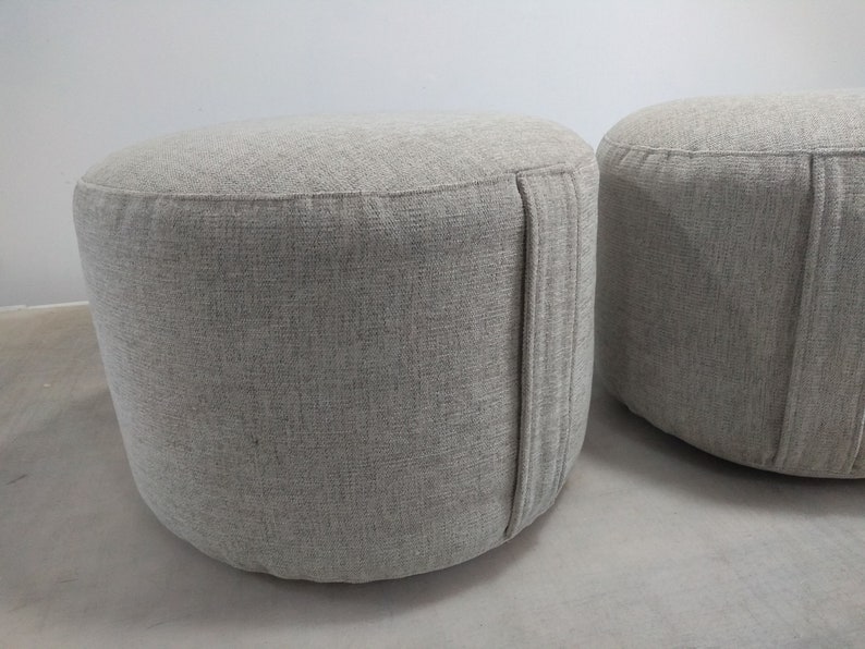 K&ouml;nnte beinhalten: Zwei runde graue Stoffhocker mit einer dezenten Textur. Die Hocker haben eine Naht an der Seite.