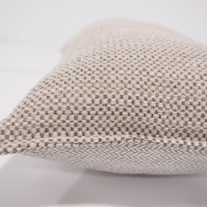 May include: A beige throw pillow with a textured woven fabric. The pillow has a subtle herringbone pattern on the bottom edge.