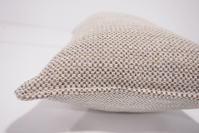 May include: A beige throw pillow with a textured woven fabric. The pillow has a subtle herringbone pattern on the bottom edge.