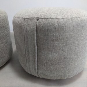 K&ouml;nnte beinhalten: Zwei graue Stoffhocker mit einer strukturierten Oberfl&auml;che. Die Hocker sind rund und haben eine Naht an der Seite.