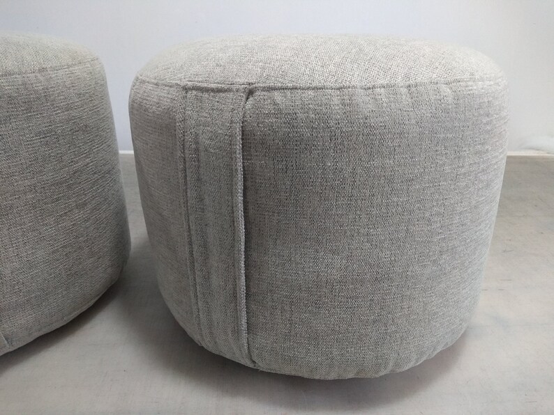 K&ouml;nnte beinhalten: Zwei graue Stoffhocker mit einer strukturierten Oberfl&auml;che. Die Hocker sind rund und haben eine Naht an der Seite.